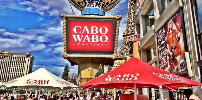 A photo taken during the daytime of Cabo Wabo Cantina on the Las Vegas Strip with a Cabo Wabo Cantina sign under the Paris's hot air balloon statue, and red and white tents that say Cabo Wabo with Caesars Palace pictured in the background