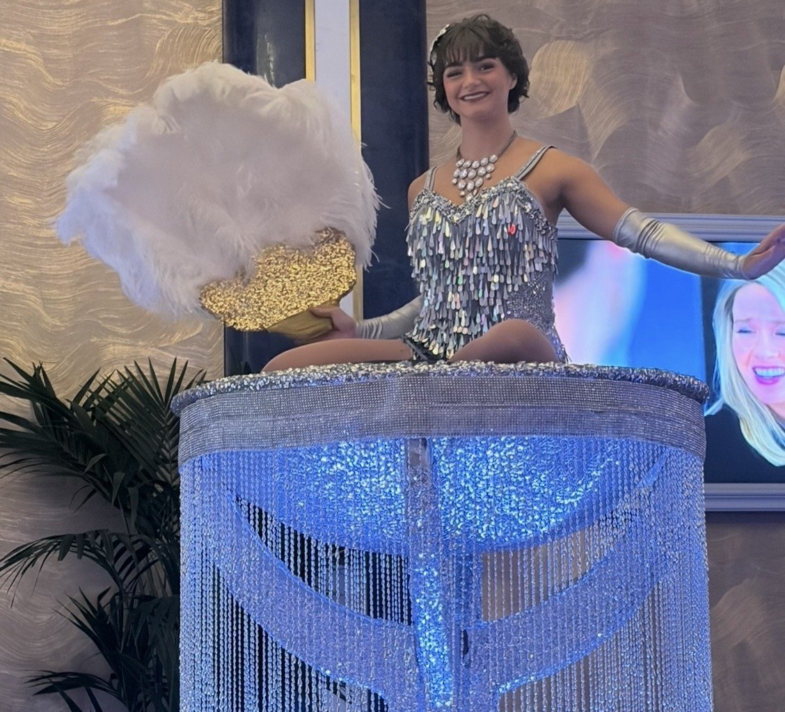 A showgirl in a silver sequined costume with a feather fan, perched atop a beaded silver platform