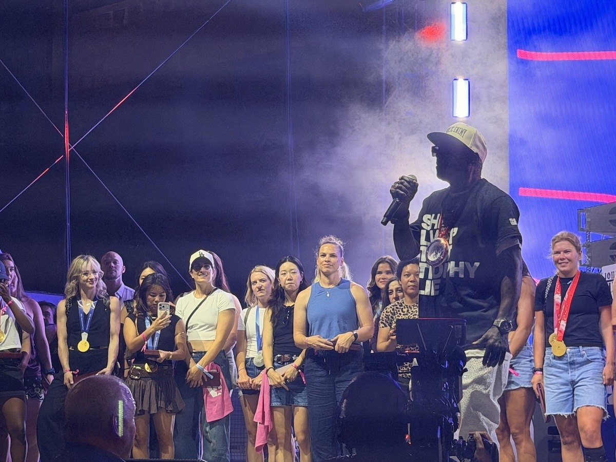 Flavor Flav stands on a stage, holding a mic in front of female Olympians from Team USA.