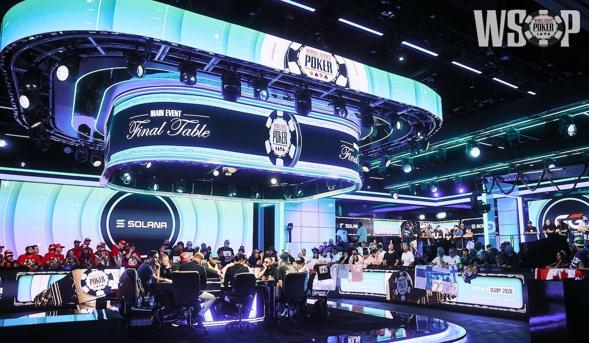 The main event final table of the World Series of poker 2026. Players sit at the final table, focused on playing poker while several fans gather around to cheer them on behind barriers with LED screens. Various screens around the image display World Series of poker imagery with poker chips and Solana imagery with blue and green backgrounds.