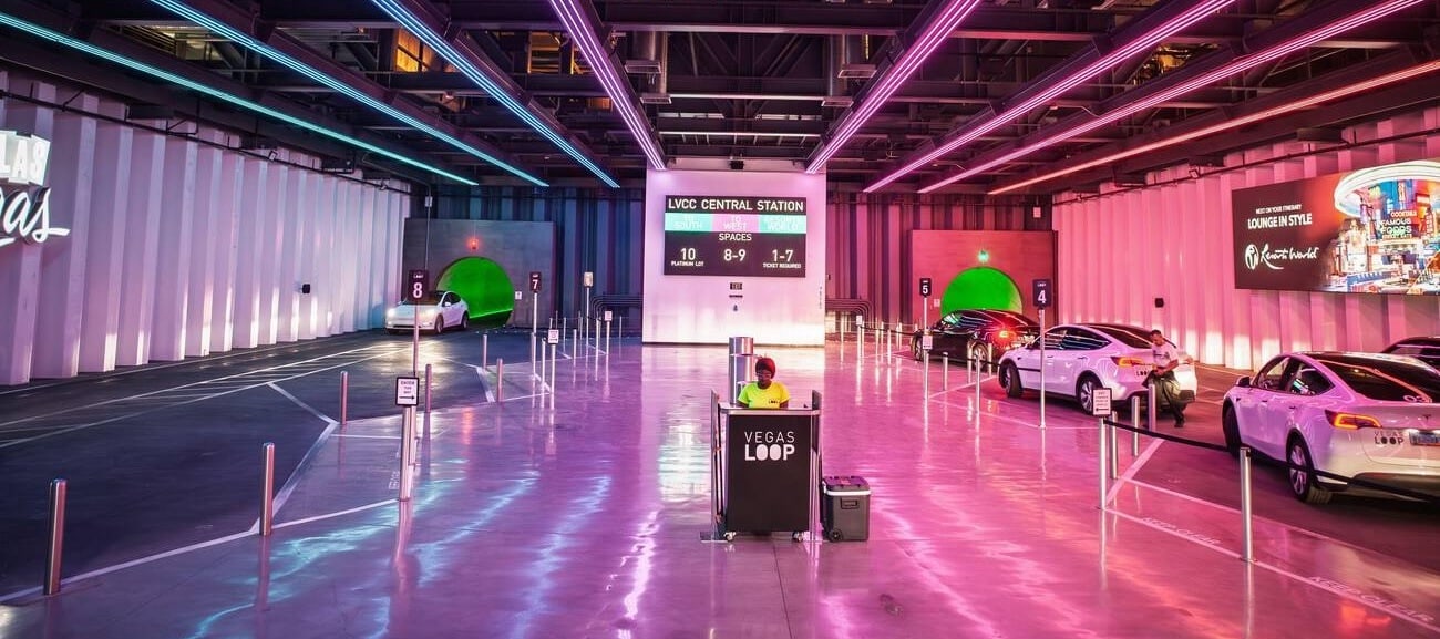 Interior of the Las Vegas Convention Center Central Station for the Las Vegas Loop. The area is lit by purple and pink lights. There is a woman in a neon green shirt standing at a podium that reads Vegas loop. Several Teslas are displayed on the right side of the screen with a Resorts World ad on the LED screen as well. The Teslas are leading in and coming out of a green tunnel Underground