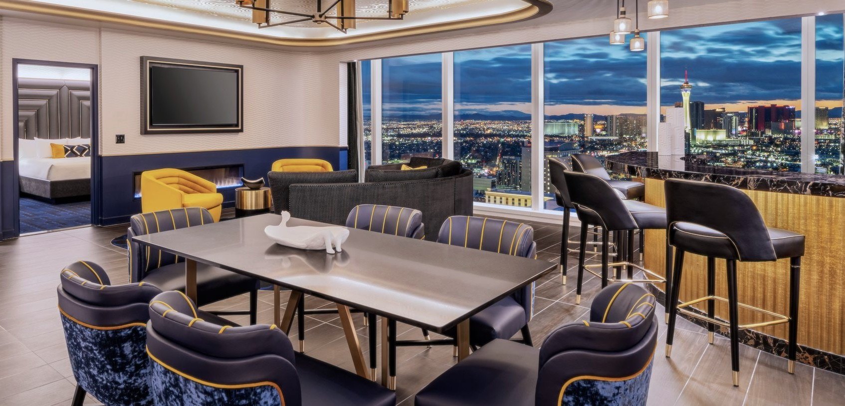 Inside of the Founder's Platinum Suite at Circa. Pictured is a dining table with several grand blue chairs, and then a mini bar a door that shows a bedroom, a TV a sofa, a view of the Las Vegas Strip at sunset.