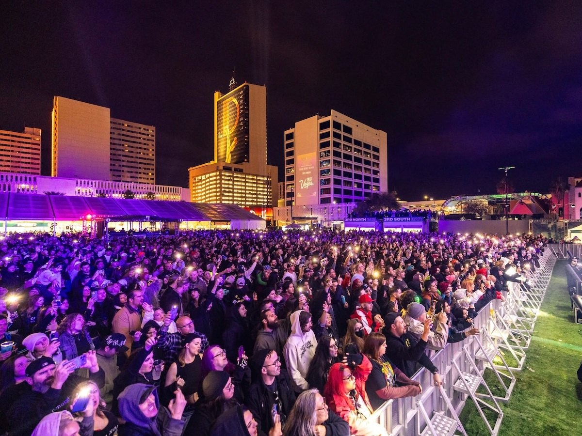 a crowd at the rails of the Downtown Las Vegas Event Center grass lawn, with purple lights and several downtown Las Vegas buildings pictured in the background.