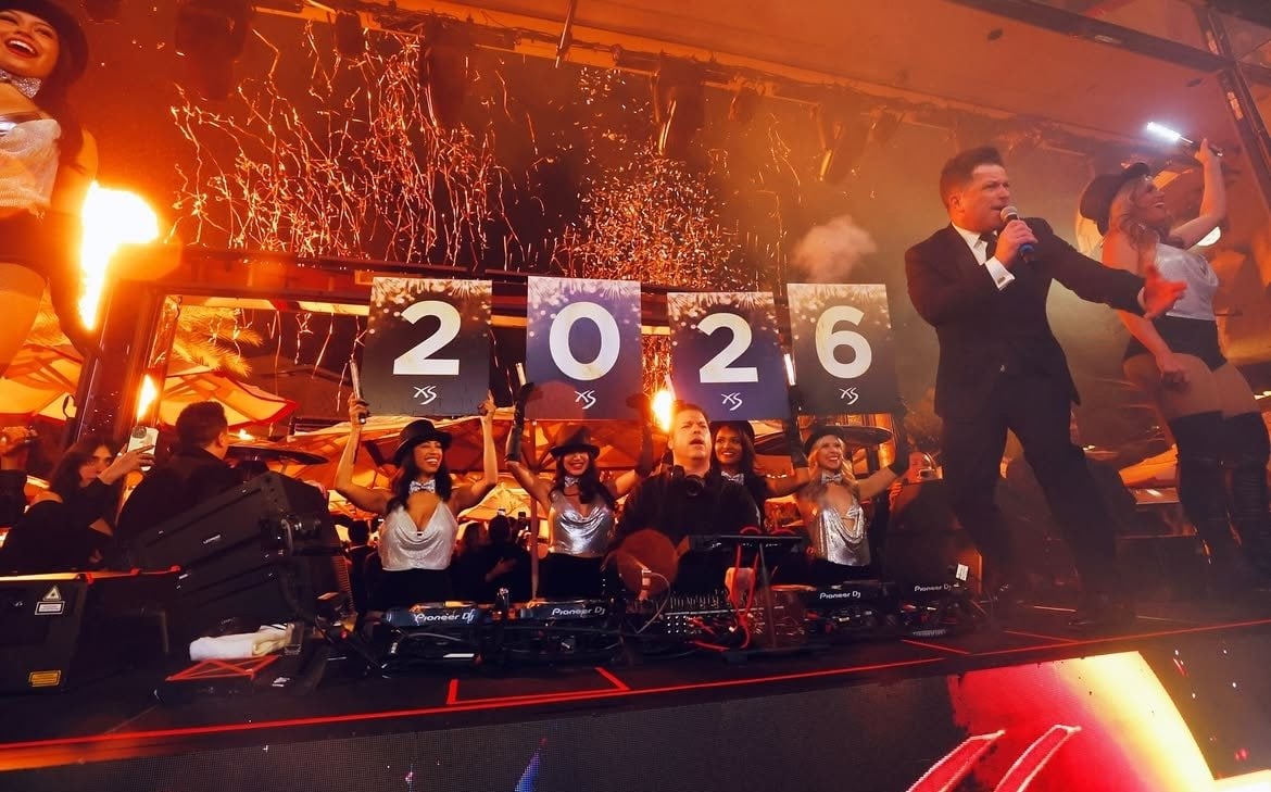 RÜFÜS DU SOL DJing at XS Nightclub on New Year's Eve in 2025. Women in silver tops and hats hold up large cards that show 2026 in a man in a suit with a microphone is standing on the stage.