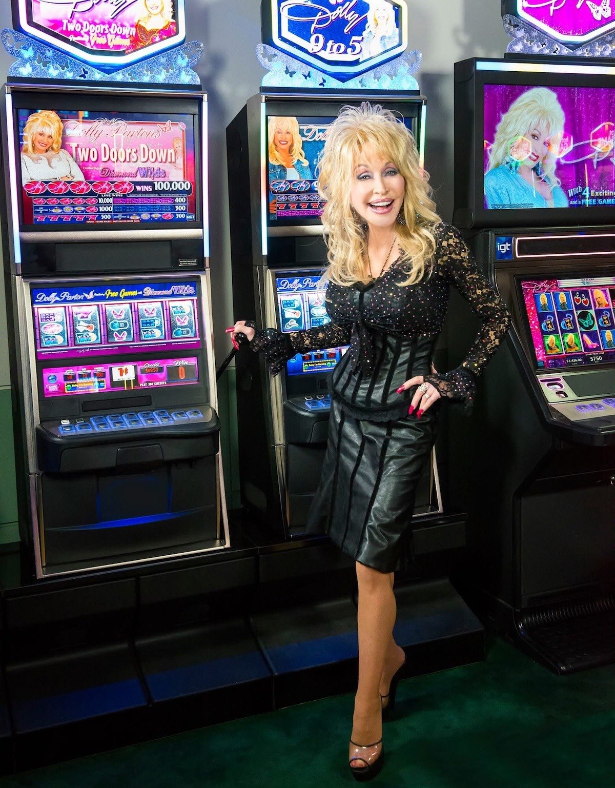 Dolly Parton wear a black dress and stands in front of branded slot machines including Dolly Parton, two doors down, and 9 to 5.
