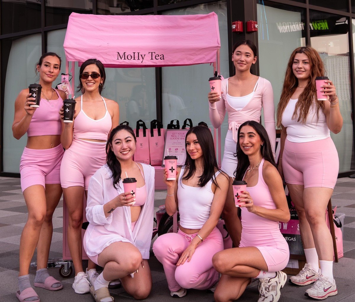 Seven women in pink and white outfit stand in front of a pink molly tea booth smiling and holding drinks