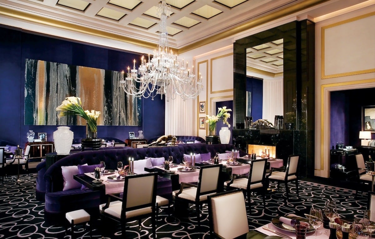 Elegant fine-dining room at Restaurant de Joël Robuchon inside MGM Grand in Las Vegas, with a curved dark-blue velvet banquette, crystal chandelier, and formally set tables.