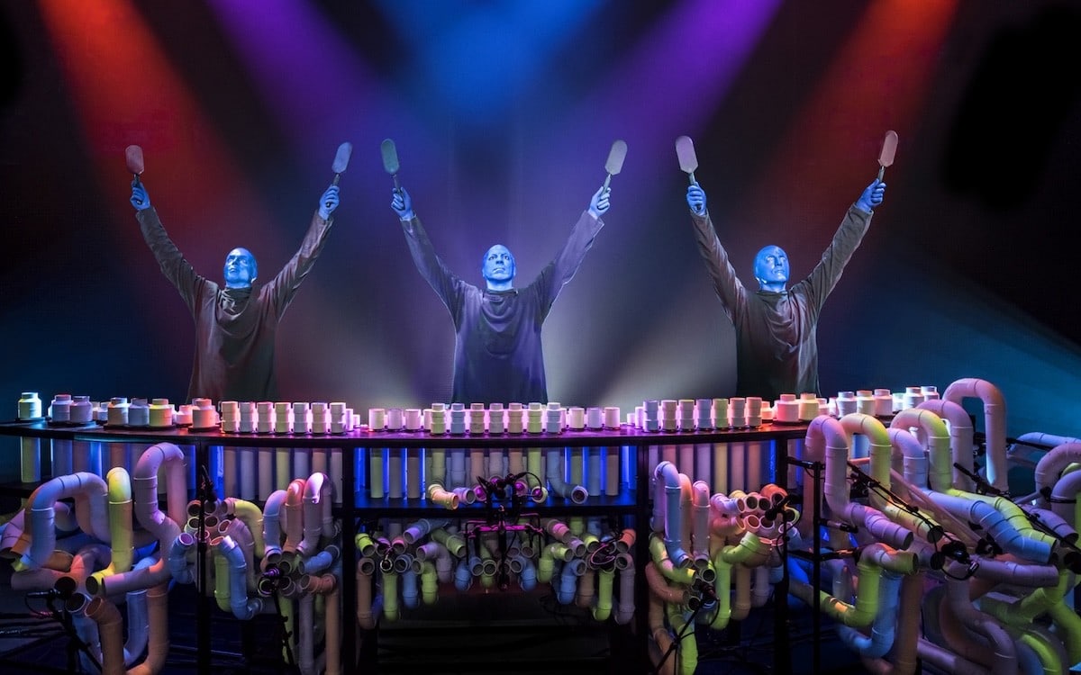 three blue-painted performers raising drumsticks over a PVC pipe instrument setup