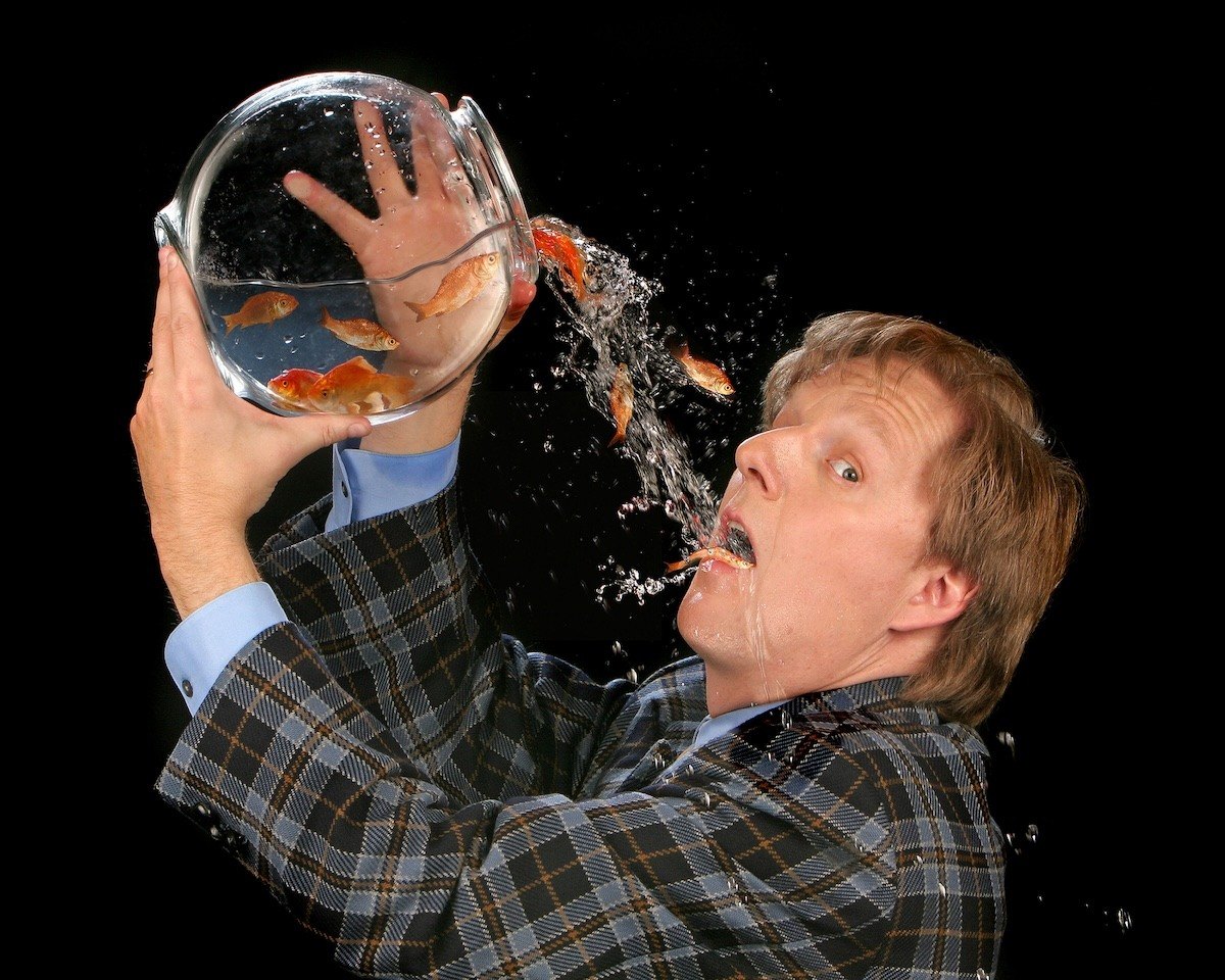comedian holding a fishbowl overhead as water and goldfish spill toward his mouth