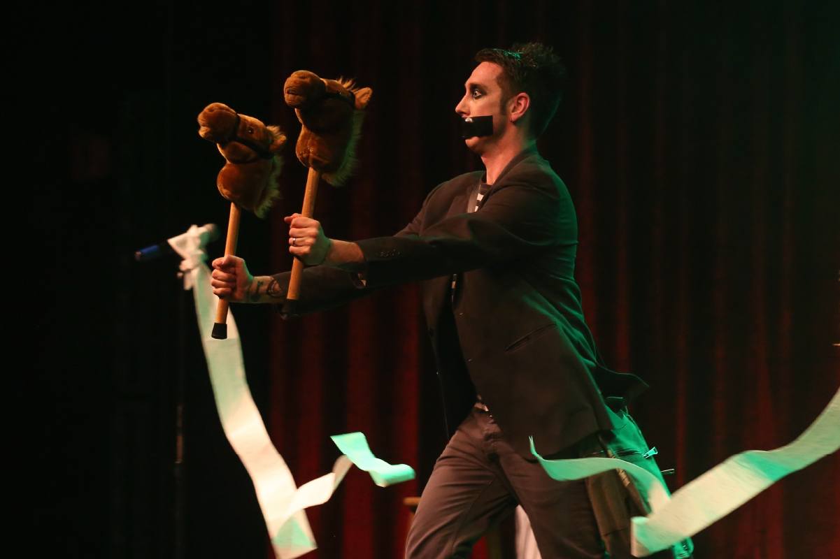Tape Face with tape over his mouth holding two hobby-horse sticks on stage
