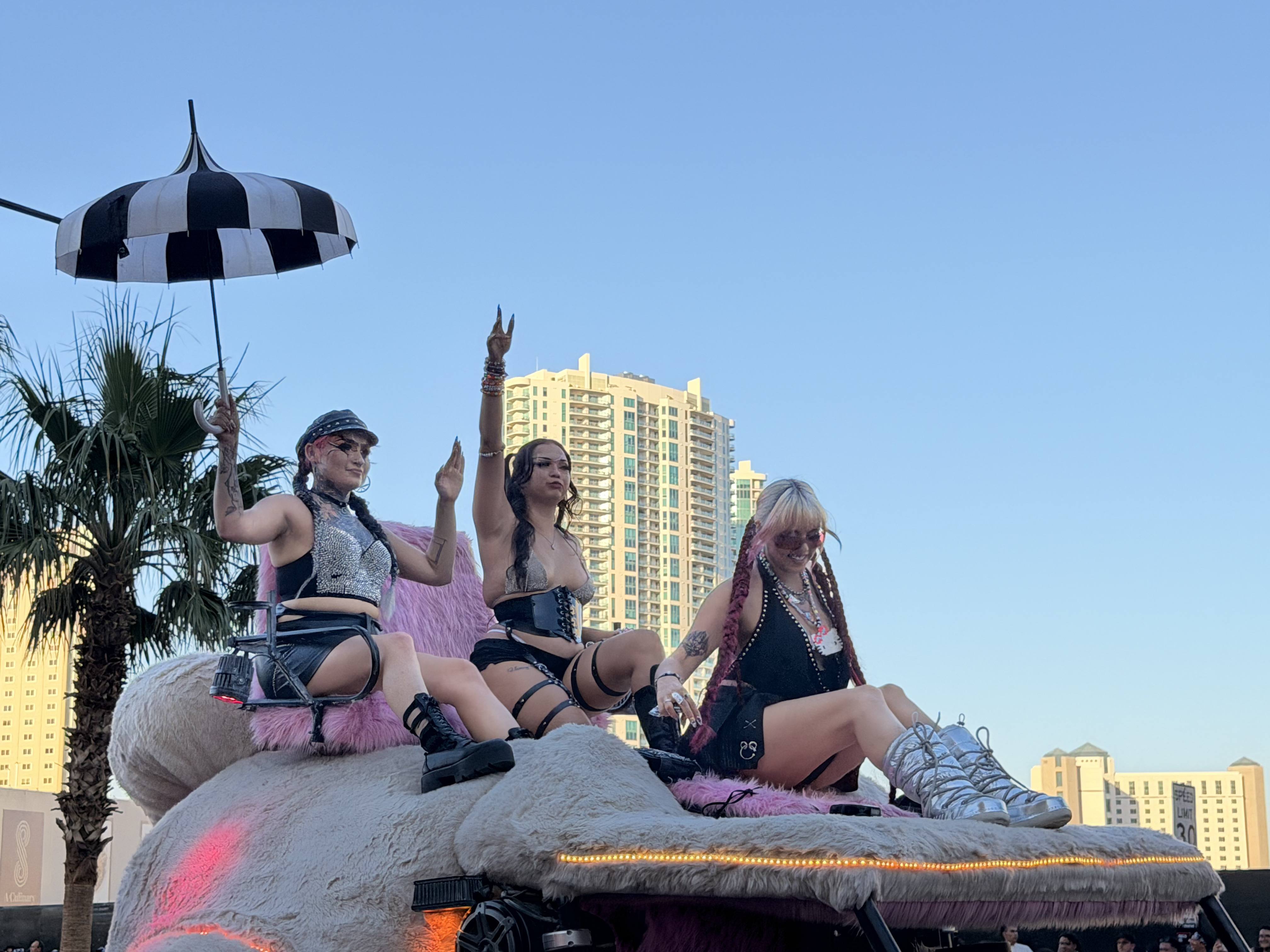 Three women in festival attire sit on a car in World Party Parade