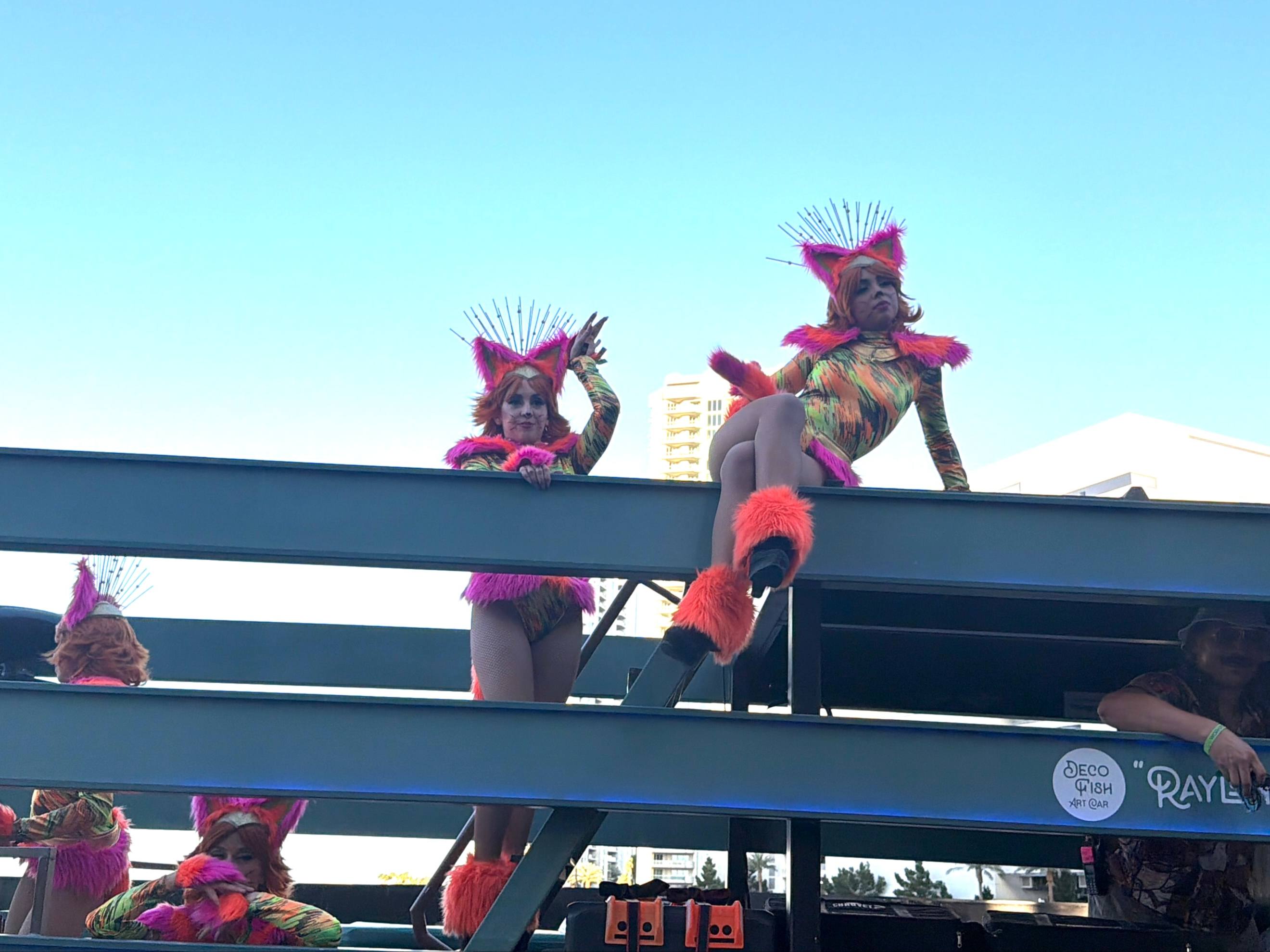 World Party Parade performers in colorful cat costumes with pink and orange fur accents and feathered headpieces posing on a Deco Fish Art Car float